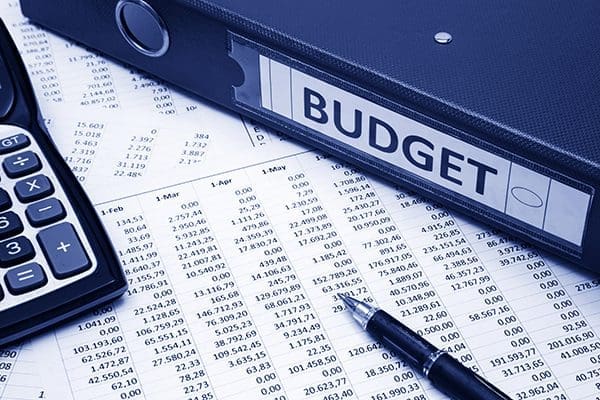 Spreadsheets with monthly budgets and a budget binder lay on a desk next to a calculator and pen.
